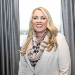 Professional headshot of Jacqueline Carver, wearing a beige blazer and plaid scarf in front of a dark grey window curtain.