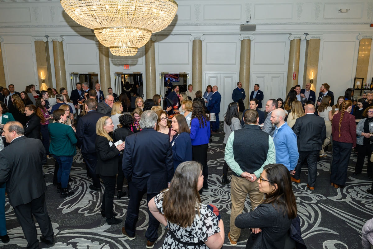 A large group of people in formal attire are mingling at an indoor event. The room is adorned with elegant chandeliers and patterned carpeting. Tall columns and white walls frame the space. Attendees are engaged in conversation, some holding drinks. Doors are visible in the background.