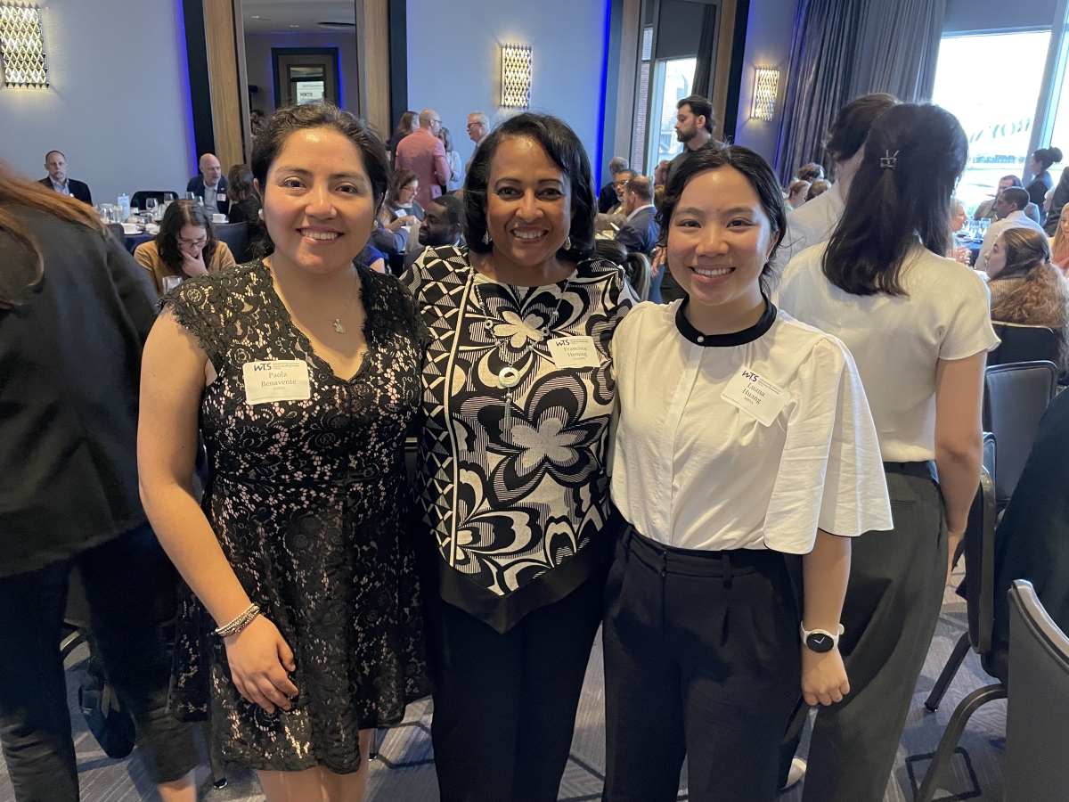 Three women stand together and smile at the camera. They are wearing business casual clothing and are posing in a room filled with tables and other people.