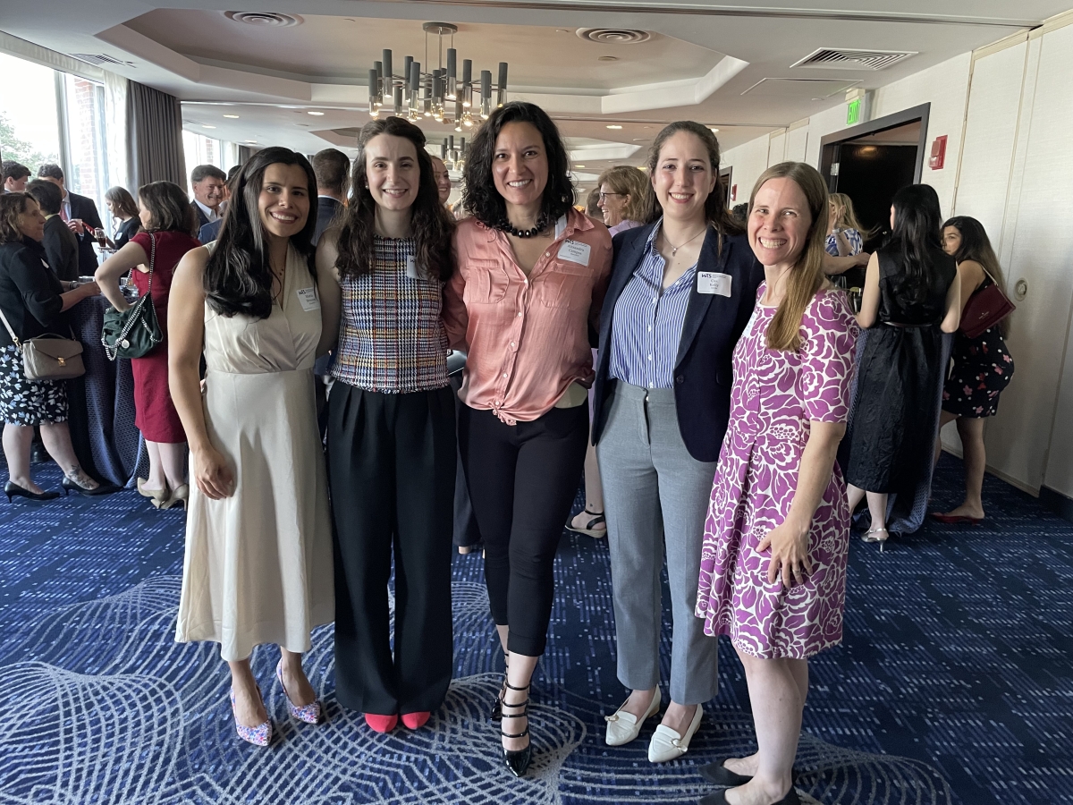 Five women wearing business casual clothing stand together and smile at the camera.