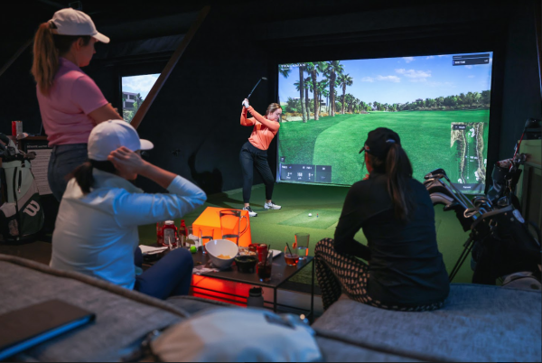 Three women in gold attire watching as a fourth woman swings a gold club in an indoor golf simulator bay at Five Iron Golf.