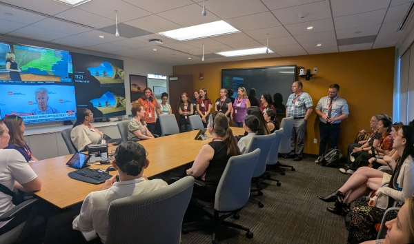 A tour group sitting and standing around a conference room while a woman presents to the group.