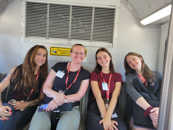Four women sitting in the backseat of a bus and smiling at the camera.
