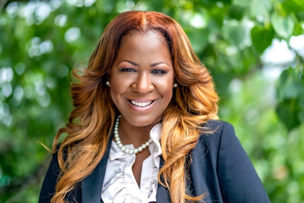 Woman smiling wearing a white blouse, navy blazer and pearl necklace in front of a blurred green foliage background