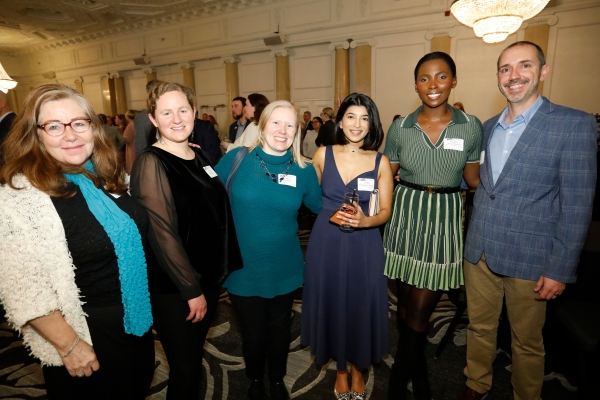 Group of six professionals networking at a formal event in a grand ballroom with ornate chandeliers. Attendees wear business attire and name tags while socializing.