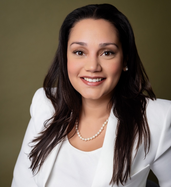 Professional head shot portrait of a woman (Victoria Palek) with long dark hair, wearing a white blazer, white top, and a pearl necklace, standing against an olive-green backdrop.