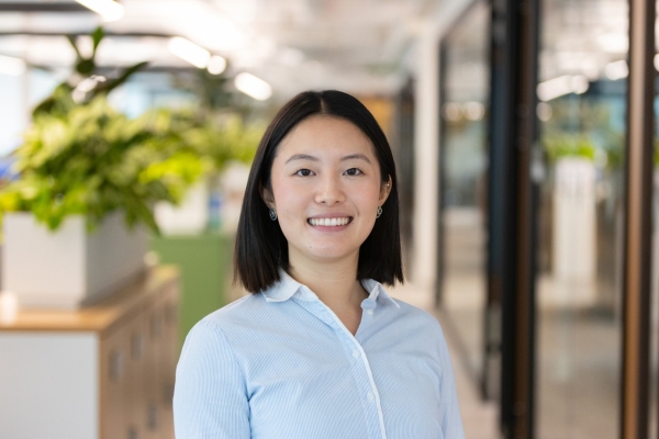The headshot shows a person with straight, medium‑length dark hair, wearing a light blue button‑down shirt, standing in a bright indoor corridor with plants in the blurred background. 