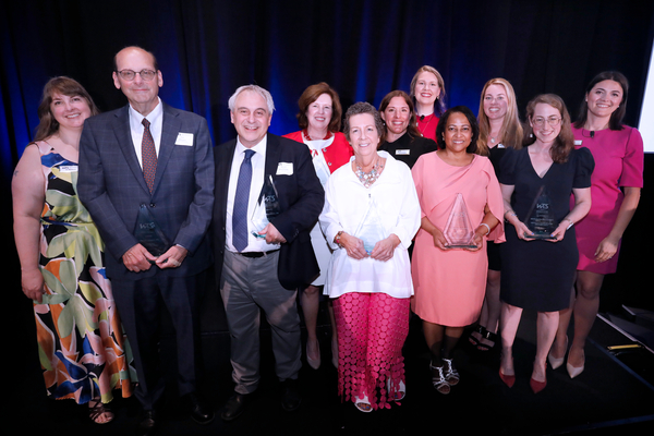 Group photo of award recipients and presenters standing on a stage in front of a dark blue backdrop. Eleven people pose in two rows, with five individuals in the front holding clear glass award trophies. Attendees are dressed in business and formal attire in a variety of colors, including white, pink, black, red, and patterned fabrics. 