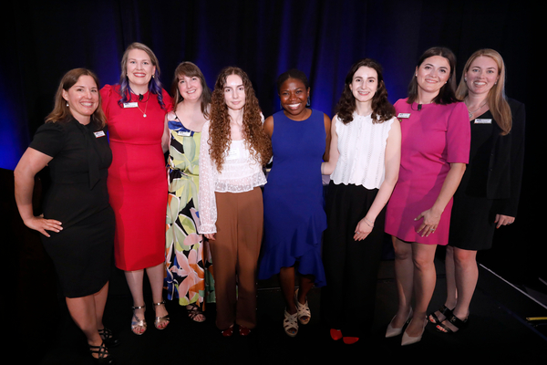 Group photo of scholarship recipients and presenters standing on a stage in front of a dark blue backdrop. Eight attendees are dressed in business and formal attire in a variety of colors, including white, pink, black, red, and patterned fabrics. 