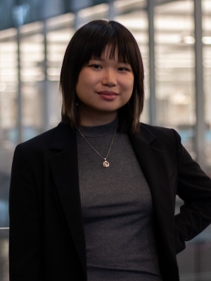 Headshot of Xinyi Looi wearing a gray shirt and black jacket in a building atrium.