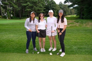 Four women golfers stand together on a lush green golf course, smiling at the camera. They are dressed in sporty golf attire, each holding a club. Tall trees line both sides of the fairway in the background, and the weather appears sunny and pleasant.