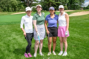A second photo of four women smiling on a golf course in golf attire and holding golf clubs.