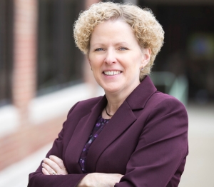 Professional headshot of a blonde woman in a burgundy suit with her arms crossed.