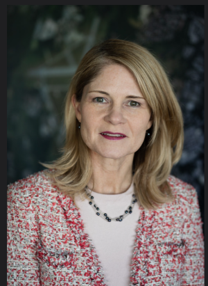 Professional headshot of a blonde woman in a multicolored suit jacket, white shirt, and necklace looking straight into the camera.