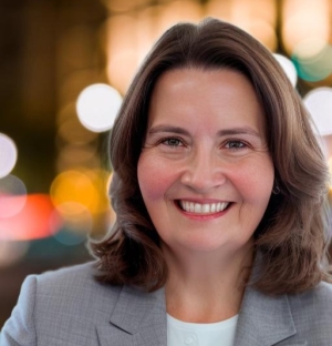 Smiling woman with shoulder-length brown hair in a light gray blazer, in front of a blurred city lights background.