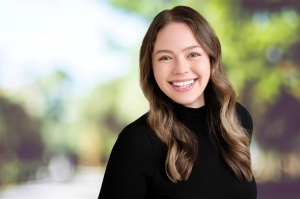 Smiling woman with long wavy brown hair in a black turtleneck, in front of a blurred outdoor background.