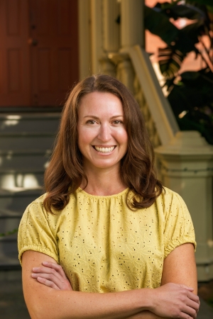 Smiling woman with long brown hair in a yellow blouse, standing with arms crossed in front of a building.