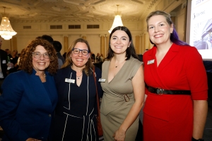 Four professional women stand together smiling at a networking event in an elegant ballroom. From left to right: woman in blue blazer, woman in navy dress with white trim, woman in beige dress, and woman in red dress with black belt.