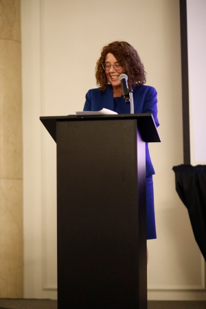 A woman with curly brown hair and glasses wearing a vibrant blue blazer speaks at a podium with a microphone, smiling as she addresses an audience at a professional event.